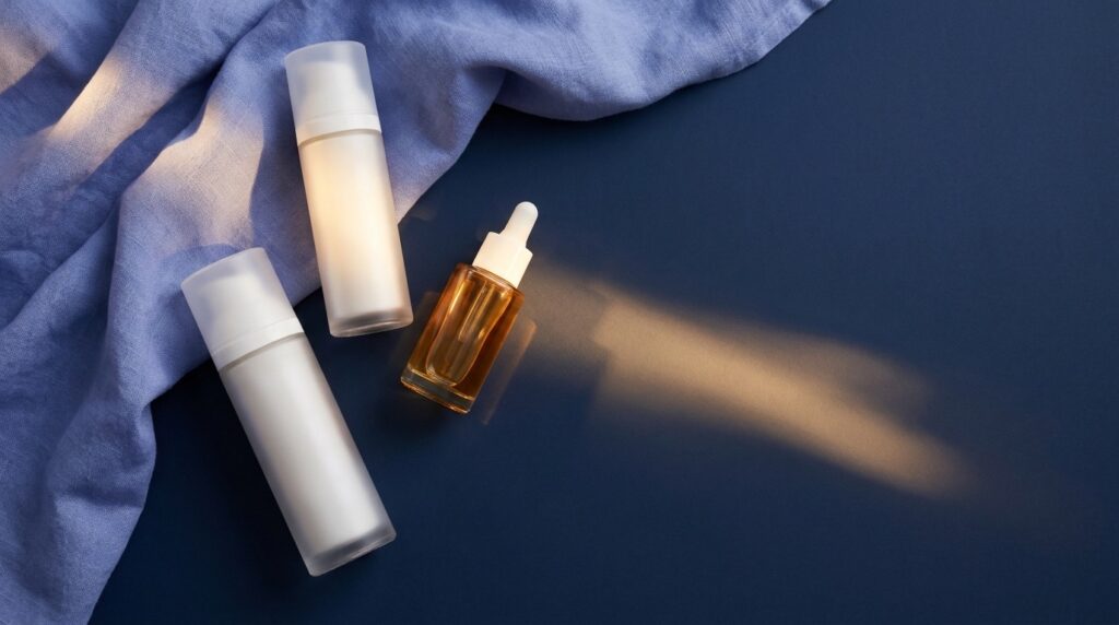 Editorial still life of unbranded frosted-glass sunscreen bottles and a sun-serum dropper on a navy surface with periwinkle linen and a streak of sunlight