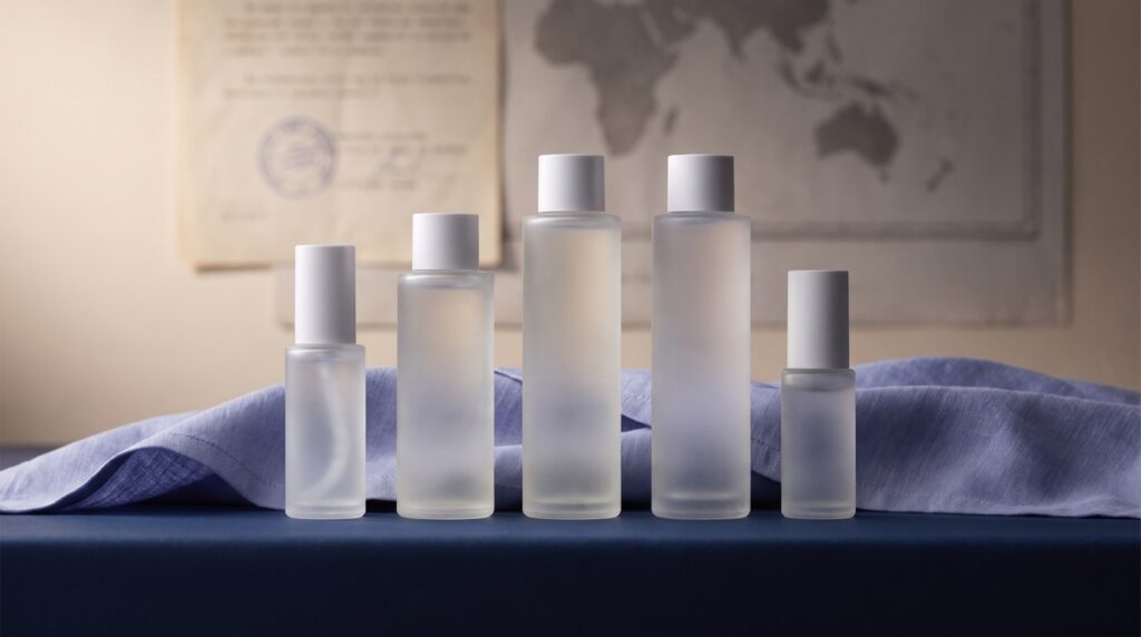 Editorial conceptual still life of a lineup of five unbranded frosted-glass Korean skincare bottles on a navy surface with periwinkle linen, a faint stamped approval document and world map blurred behind